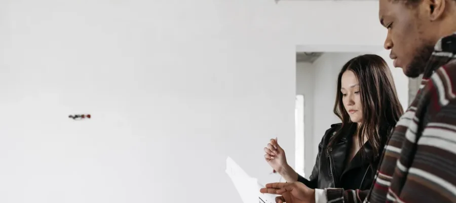 A couple discusses home renovation ideas in an empty room with white walls.