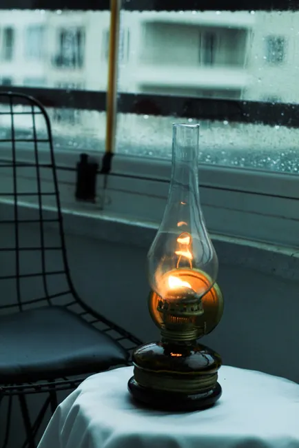 A glowing kerosene lamp on a table by a rainy window creating a cozy, vintage atmosphere.