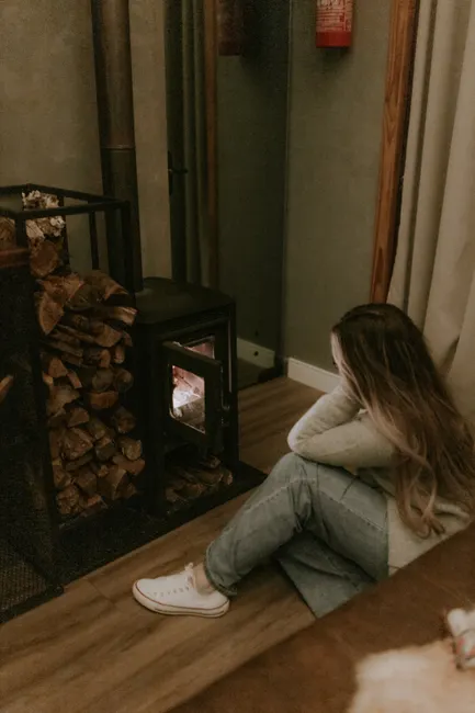 A woman sits cozily by a wood-burning stove in a relaxing indoor setting.