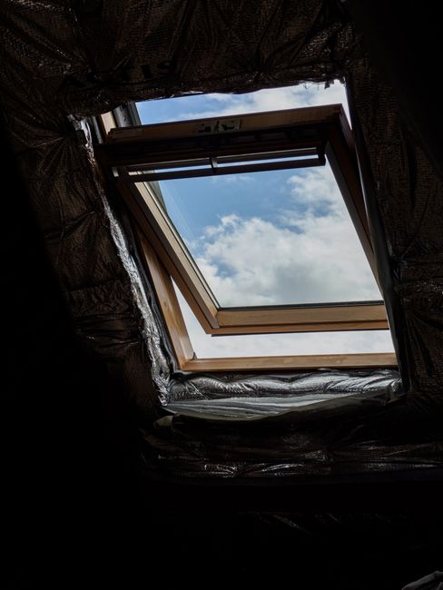 A wooden attic window opens to reveal a clear blue sky and fluffy white clouds.