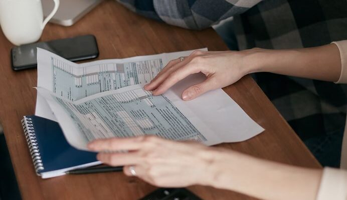 Close-up of hands organizing tax forms on a desk with a calculator, laptop, and notebook.