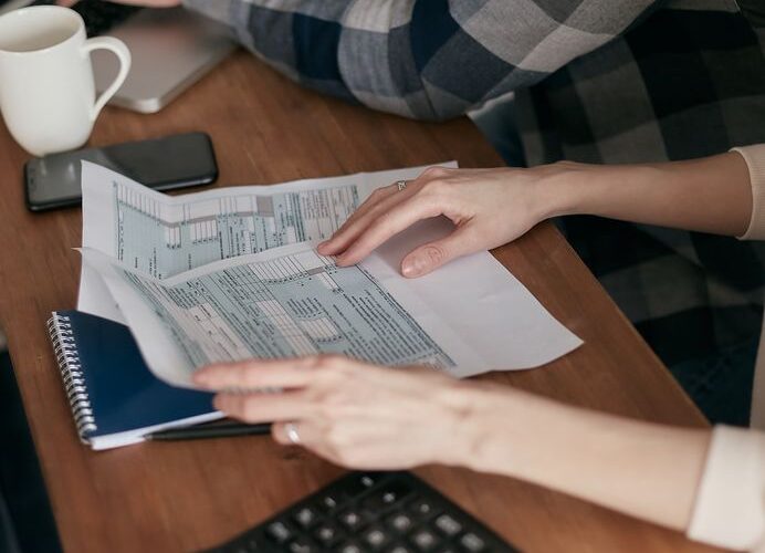 Close-up of hands organizing tax forms on a desk with a calculator, laptop, and notebook.