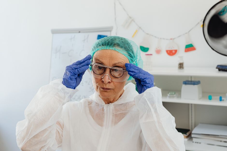 Elderly woman scientist adjusting goggles while wearing full protective lab attire including gloves and gown.