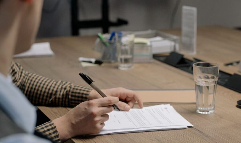 Person in business attire signing a document at a wooden table in an office setting.