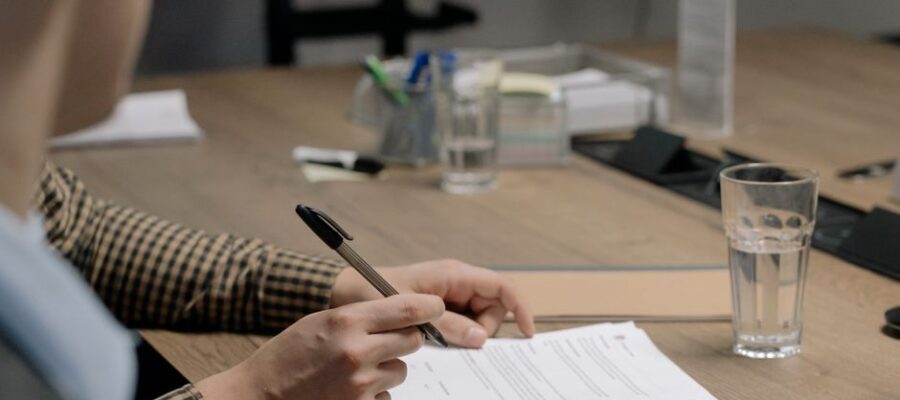 Person in business attire signing a document at a wooden table in an office setting.