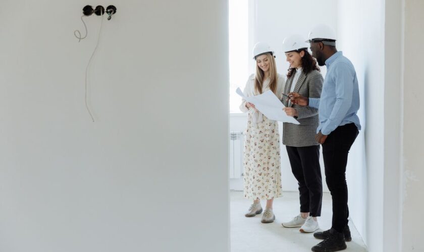 Three architects with hard hats reviewing building plans in an unfinished room.