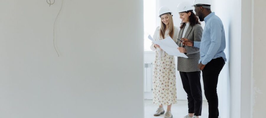 Three architects with hard hats reviewing building plans in an unfinished room.