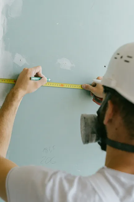 A builder in a hard hat measures a wall's dimensions with a tape measure.