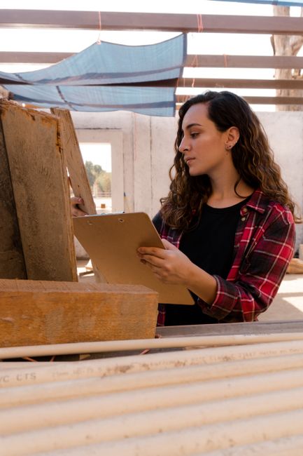 A female carpenter inspects wooden planks with a clipboard at a construction site. Professional and focused.