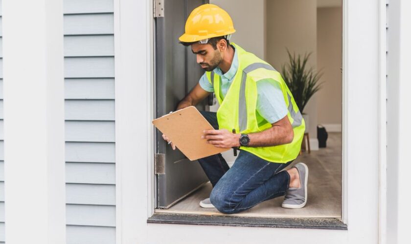 A home inspector wearing safety gear examines a house interior for safety compliance.