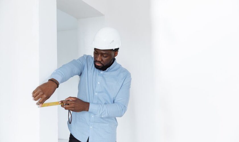 A man in a hard hat measures a wall, showcasing precision in a building interior.