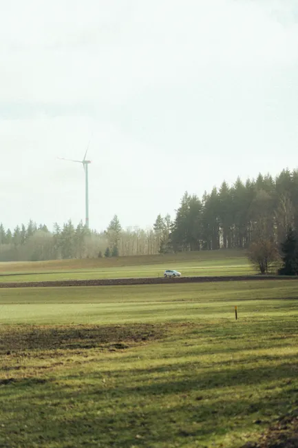 A peaceful rural scene featuring a wind turbine, coniferous forest, and a car on a road.