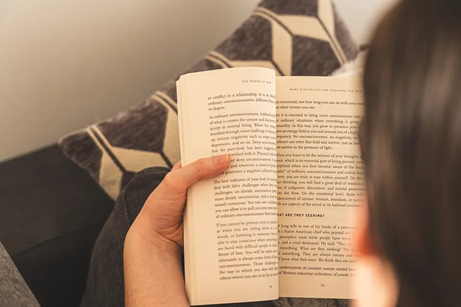 A person enjoys a relaxing reading session indoors, holding an open book on a comfy couch.