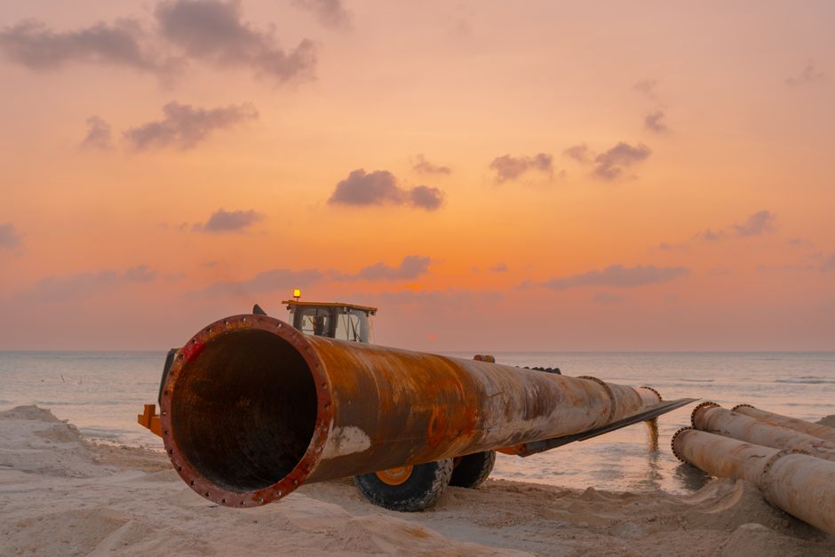 A scenic beach construction site at sunset with heavy machinery and large pipes.
