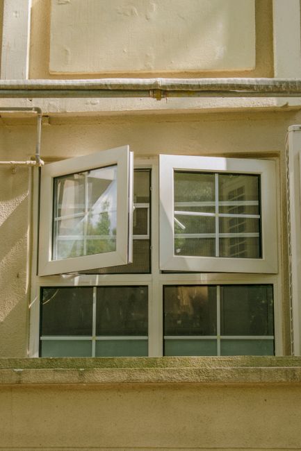 A tilted open window on a beige urban building facade with sunlight and shadows.