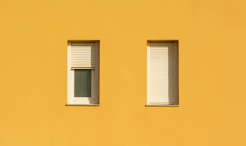 A yellow wall featuring two closed windows with roller shutters, showcasing a minimalist design.