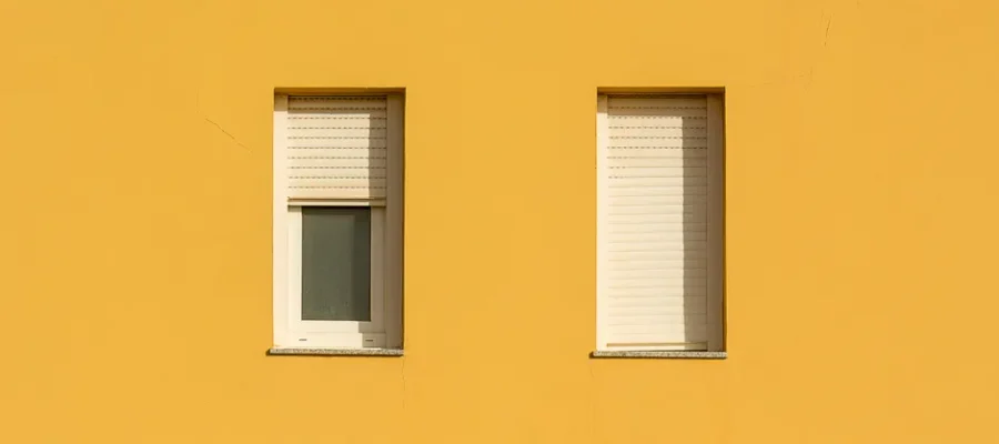 A yellow wall featuring two closed windows with roller shutters, showcasing a minimalist design.
