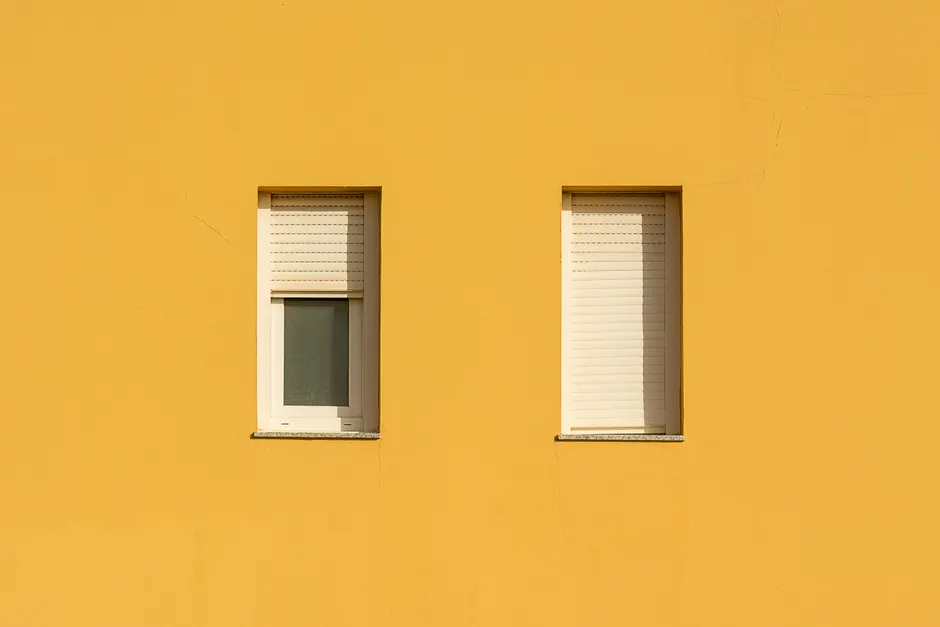 A yellow wall featuring two closed windows with roller shutters, showcasing a minimalist design.