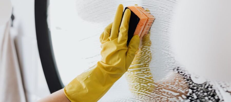 Anonymous person in yellow rubber gloves using simple sponge for cleaning mirror in bathroom