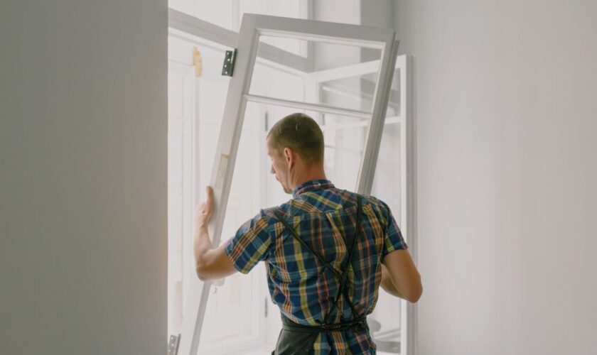 Back view of anonymous male fitter in checkered shirt and apron mounting window with hinges near white walls at home