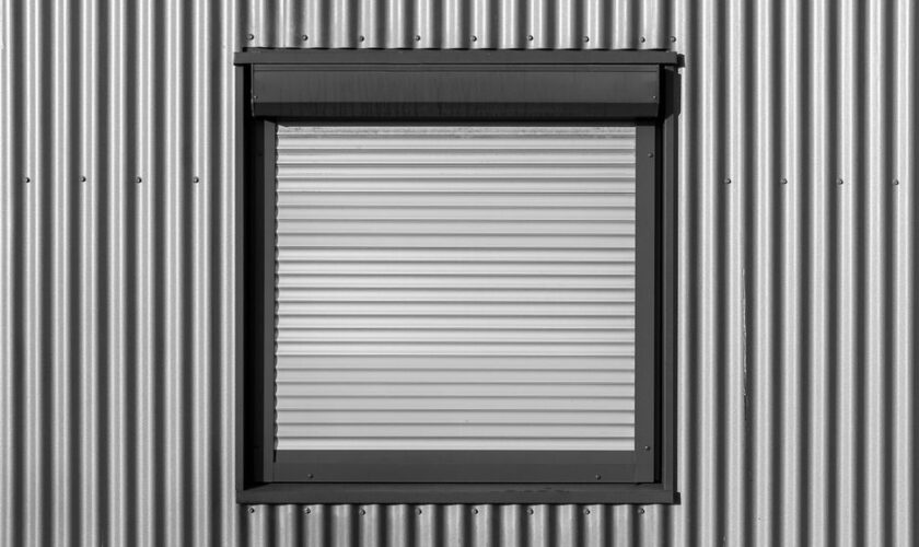 Black and white photo of a window with roller shutter on a corrugated metal wall, showcasing modern architecture.