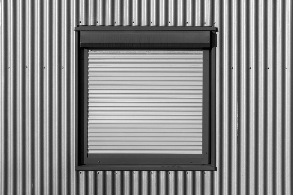 Black and white photo of a window with roller shutter on a corrugated metal wall, showcasing modern architecture.