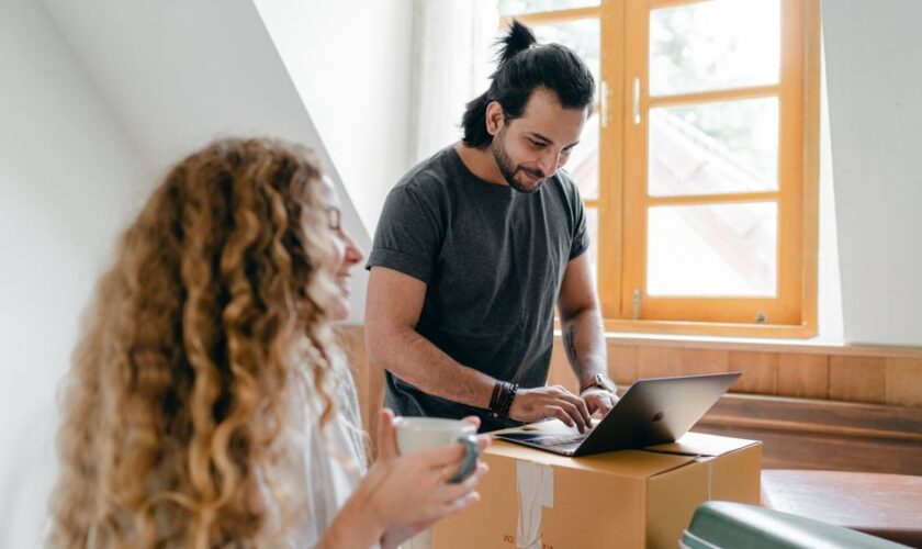 Cheerful young couple in casual clothes browsing modern netbook and having cup of hot drink while packing stuff for moving out into new house
