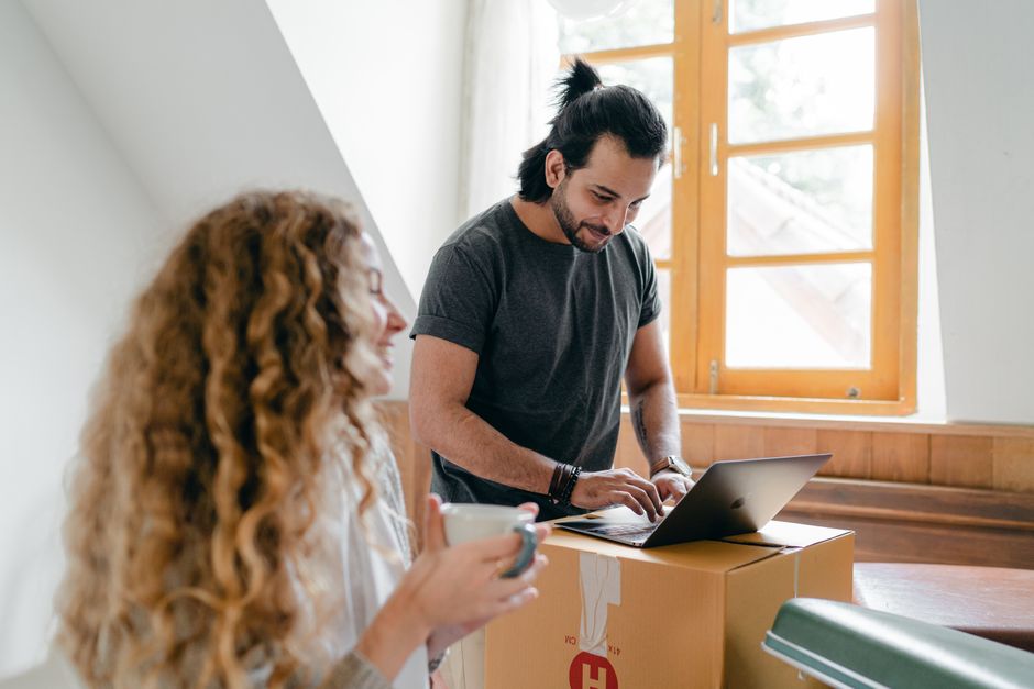 Cheerful young couple in casual clothes browsing modern netbook and having cup of hot drink while packing stuff for moving out into new house