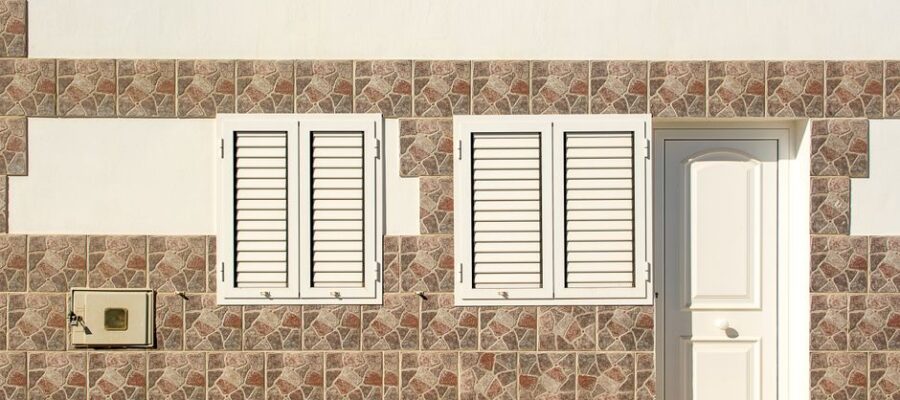 Close-up of a building facade featuring two white shuttered windows and a white door set within a mosaic tile wall.