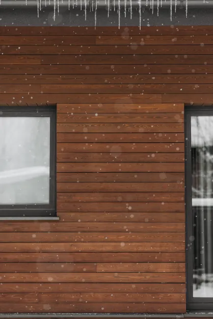 Close-up of a wooden house wall with snowflakes falling and icicles hanging.