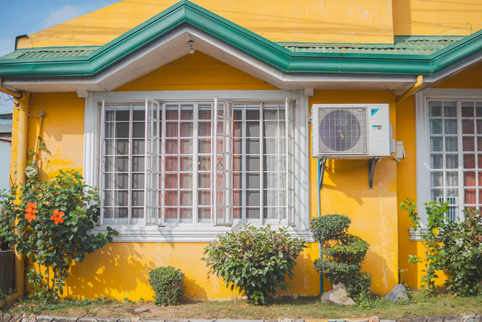 Colorful yellow house exterior with green roof, plants and air conditioner unit.