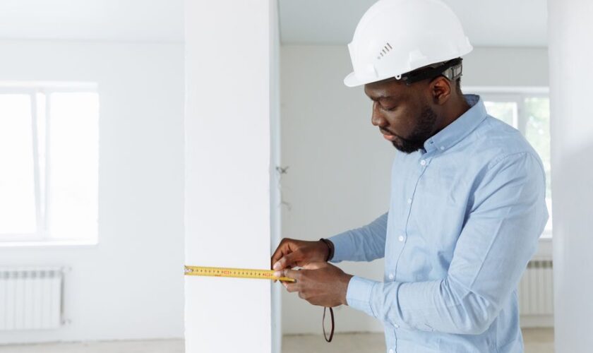 Construction worker measuring a wall indoors, wearing a white hard hat and blue shirt.