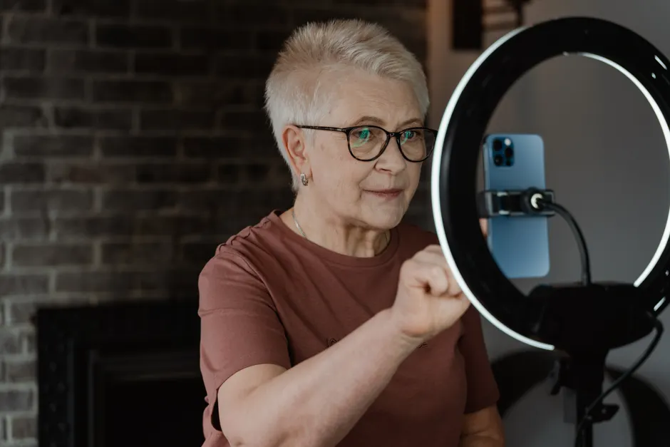 Elderly woman engaging with technology using a smartphone and ring light indoors.