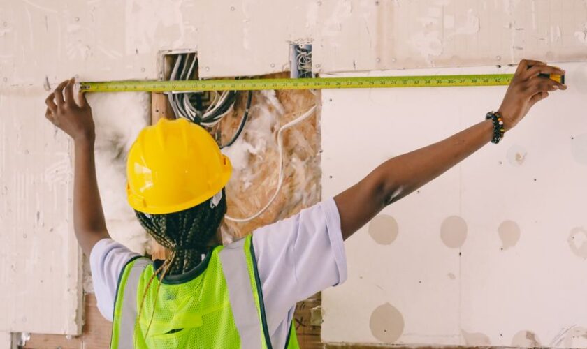 Female construction worker with hardhat and vest measuring an interior wall.