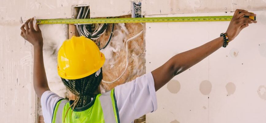 Female construction worker with hardhat and vest measuring an interior wall.