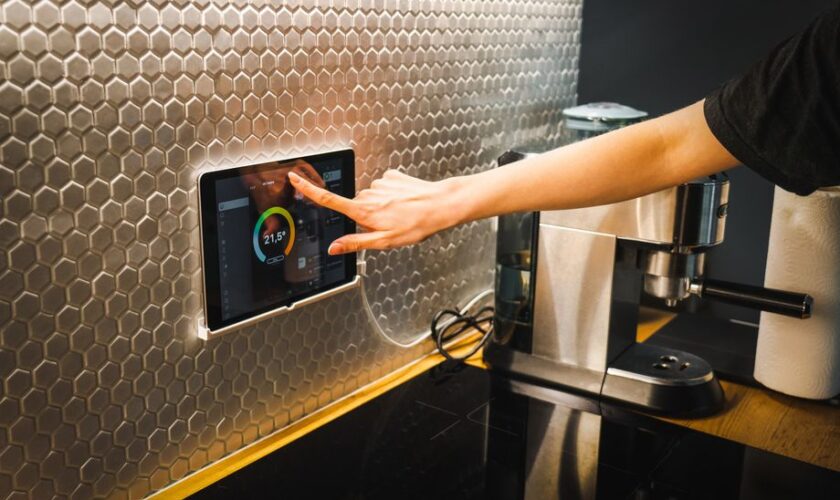 Modern kitchen scene featuring a hand using a tablet to control a smart home system.