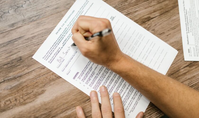 Person filling out a tax questionnaire with a pen, showing close-up on wooden table.
