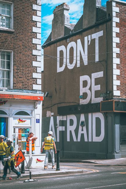 Street scene with bold 'Don't Be Afraid' mural, construction workers, and vintage architecture.