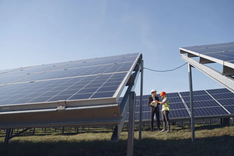 Supervisor in hardhat and formal wear discussing project with workman in uniform while standing with papers near modern solar panels under blue sky