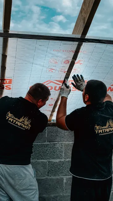 Two construction workers collaborate to install roof insulation sheets under a clear sky.