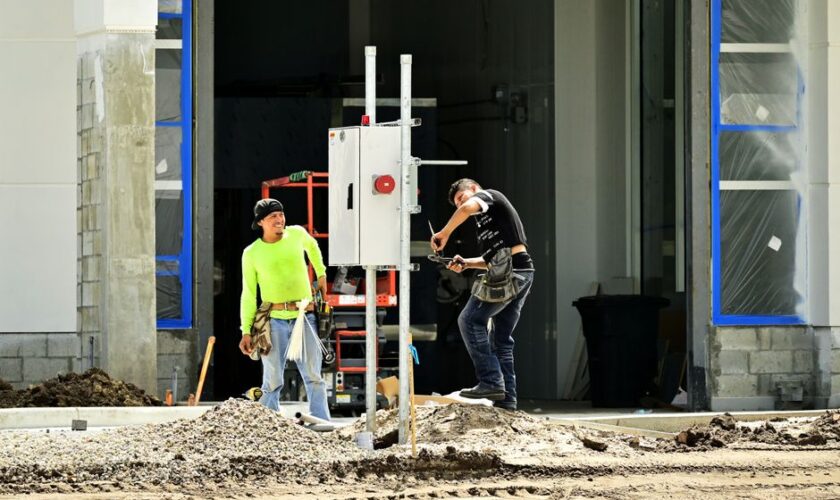 Two construction workers install an electrical panel outside a building site, wearing safety gear.