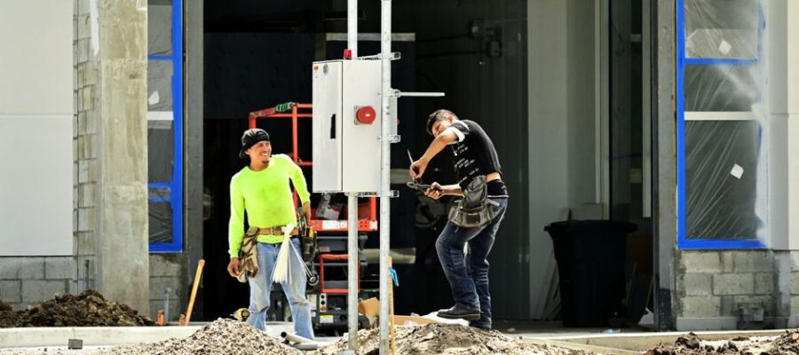 Two construction workers install an electrical panel outside a building site, wearing safety gear.