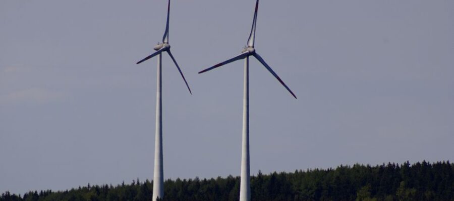 Two wind turbines stand tall against a backdrop of lush German forest under a clear sky.