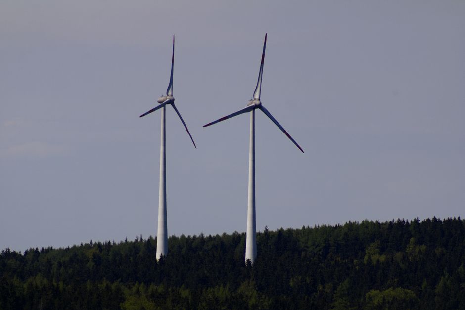 Two wind turbines stand tall against a backdrop of lush German forest under a clear sky.