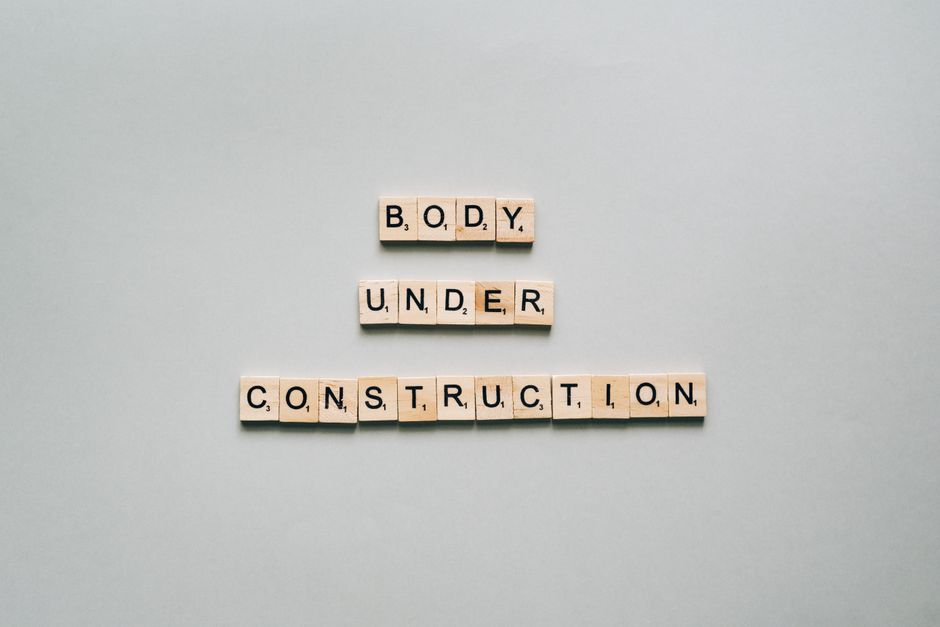 Wooden scrabble tiles spell 'Body Under Construction' on a minimalist white background, conveying motivation.