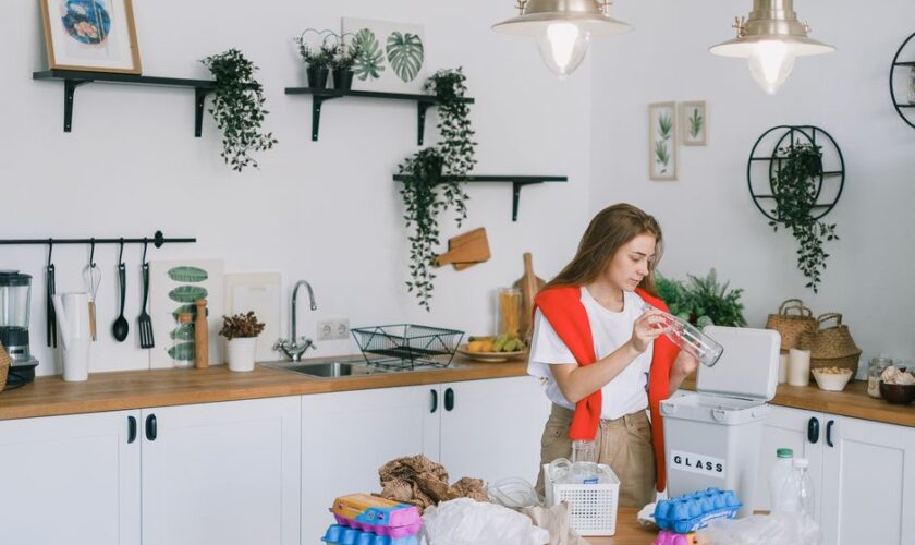 Young female in casual clothes standing in light modern kitchen with shelves and cabinets while sorting plastic glass and paper wastes