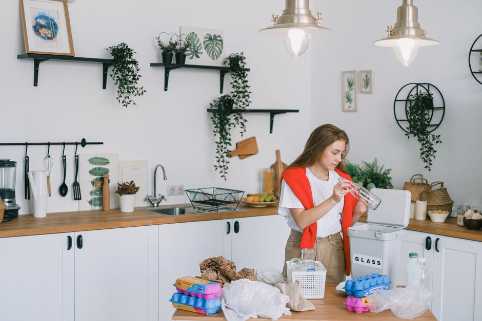 Young female in casual clothes standing in light modern kitchen with shelves and cabinets while sorting plastic glass and paper wastes