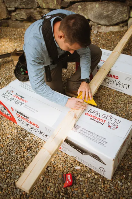 A construction worker carefully measures a piece of lumber for roofing work outdoors.