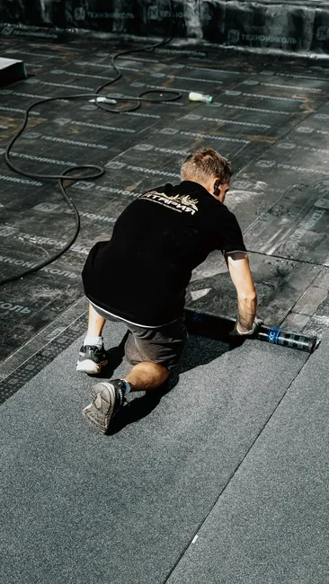 A construction worker installs asphalt roofing material under the sun, demonstrating professional craftsmanship.