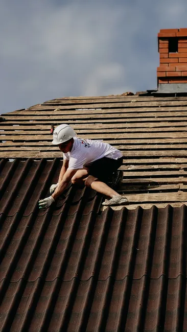 A construction worker installs roofing tiles wearing protective gear on a sloped rooftop.